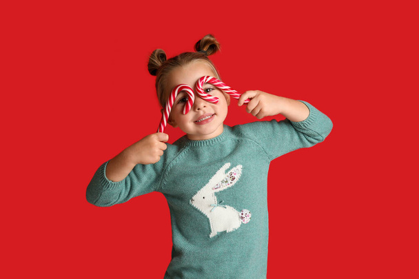 Cute little girl in knitted sweater holding candy canes on red background. Celebrating Christmas - Foto, afbeelding
