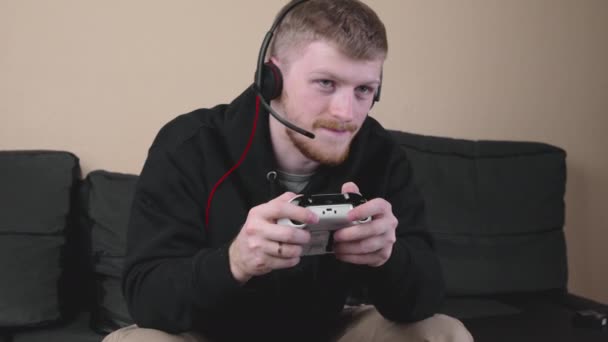 Молодой белый мужчина в черной толстовке играет в видеоигры с геймпадом. - Кадры, видео