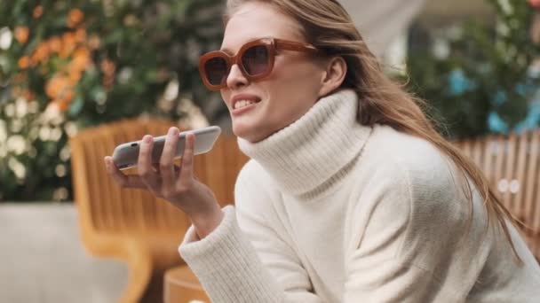 美しいですスタイリッシュな女の子でサングラスを身に着けています居心地の良い白いセーターに身を包んだストリートカフェでスマートフォンを使用して親友に音声メッセージを送信 - 映像、動画