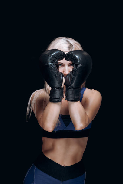A sporty blonde in boxing gloves holds her hands in front of her face smiles and prepares for training isolated on black background In the shape of a heart. - Photo, image