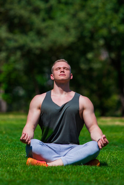 Young muscular man is doing yoga in a summer park. Sporty man is sitting in the Lotus position and relaxing outdoors - Foto, afbeelding