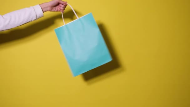 Winkelen of verkoop concept. Vrouwelijke hand houdt blauwe boodschappentas geïsoleerd op gele achtergrond. Videoband met kopieerruimte. Resolutie 4k - Video