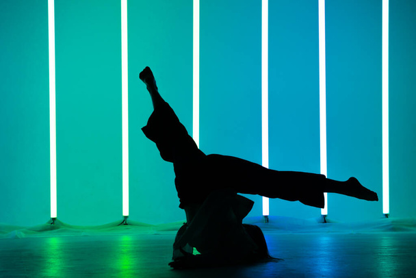 Silhouette of young woman street dancer dancing in a studio with neon lighting tube doing gymnastic exercises. Dance and music poster design. - Фото, изображение