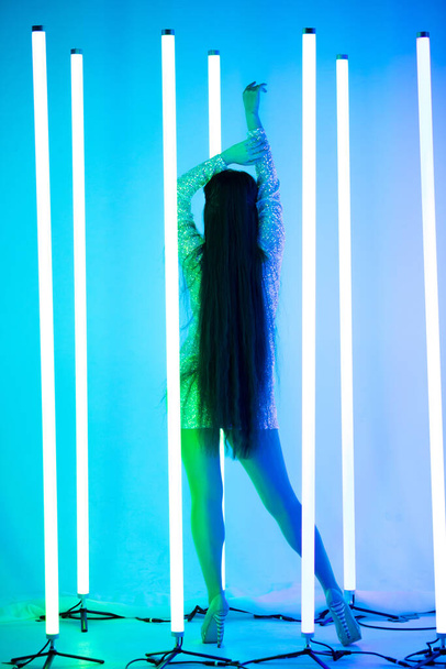 Οπίσθια όψη μιας σαγηνευτικής μελαχρινής γυναίκας με μακριά μαλλιά σε ένα κοντό γυαλιστερό φόρεμα στέκεται πάνω σε ένα φόντο από σωλήνες νέον. - Φωτογραφία, εικόνα