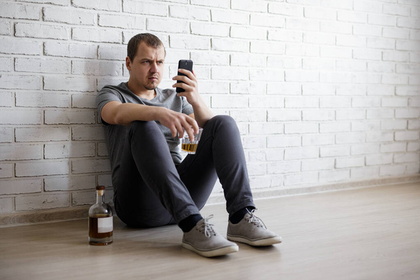 alkoholismi ja masennus käsite - nuori stressaantunut mies istuu lattialla, juo alkoholia ja käyttää älypuhelinta - Valokuva, kuva