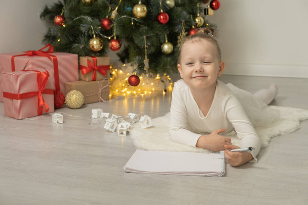 Cute little blondynka dziewczyna leżąca na podłodze w pobliżu choinki pisząc list do Świętego Mikołaja. - Zdjęcie, obraz