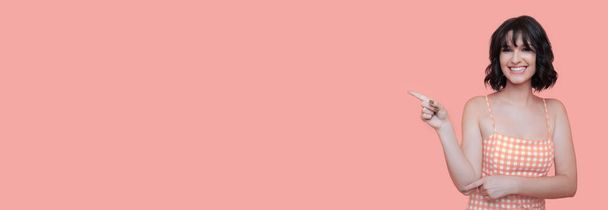 Mujer morena en ropa de verano está apuntando a una pared de estudio rosa con espacio libre mientras sonríe a la cámara - Foto, imagen