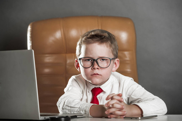 Takım elbiseli bir çocuk ve büyük patron gibi gözlüklü. Modern ofis geçmişinde bir çocuk işadamının portresi. Başarılı olmak çocuk hayalidir. Bilgisayarlı, beyaz gömlekli ve kırmızı kravatlı okul çocuğu. - Fotoğraf, Görsel