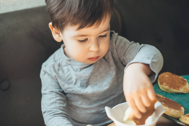 Caucasian brunette boy is eating pancakes in the kitchen sitting in an armchair - 写真・画像