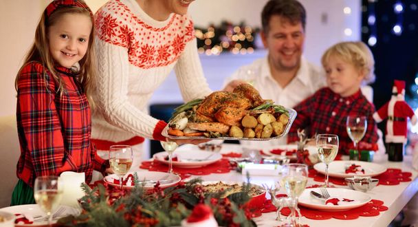 Οικογένεια με παιδιά που τρώνε γαλοπούλα Χριστουγεννιάτικο δείπνο στο τζάκι και διακοσμημένο χριστουγεννιάτικο δέντρο. Γονείς, παππούδες και παιδιά σε εορταστικό γεύμα. Χειμερινές γιορτές και φαγητό. Τα παιδιά ανοίγουν δώρα. - Φωτογραφία, εικόνα