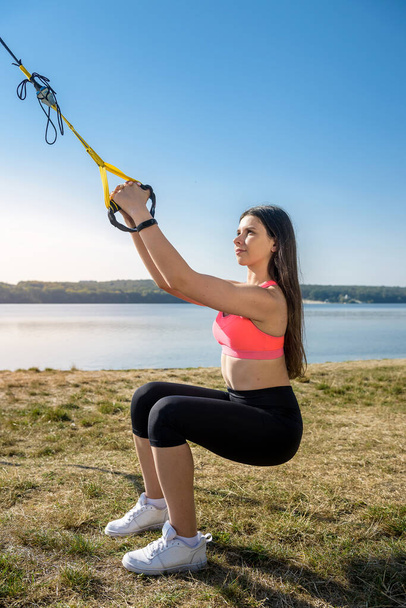 Young woman exercising with suspension trainer sling in park, near the lake. - Fotó, kép