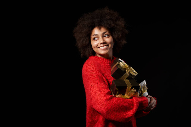 Black Friday sale, gift, present, celebration concept. Happy afro-american female holding a heap of black gift boxes with golden bows over black background, looking at blank copy space - Fotoğraf, Görsel