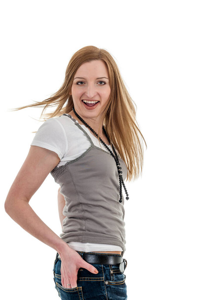 Lateral three-quarter portrait of a peppy young woman in jeans, her right hand in her hip pocket, white t-shirt and gray top against a white background, turning into the camera with flowing hair, casually turned towards the camera, smiling friendly. - Photo, Image