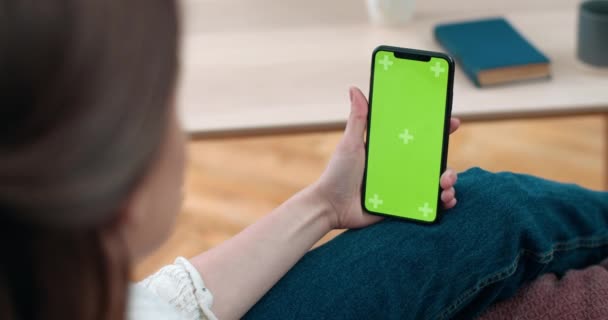 Backside view of woman holding and looking at smartphone green screen. Deaf female person having video call while sitting on sofa at home. Concept of hearing loss. - Footage, Video
