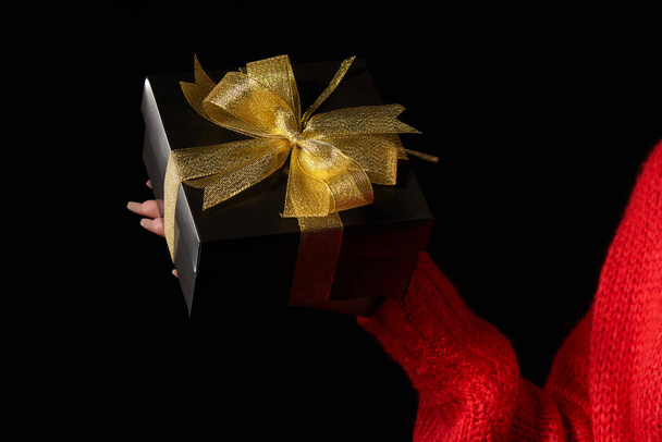 Zwarte vrijdag uitverkoop, cadeau, cadeau, feestconcept. Vrouwelijke hand met een zwart kado doosje met gouden strik op een palm, over zwarte achtergrond - Foto, afbeelding