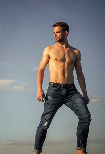 Muscular bare torso. Six packs muscular chest. Man outdoors blue sky background. Summer trends. Male beauty concept. Hot day. Athletic handsome macho wear denim pants. Muscular body. Fitness model - Foto, imagen