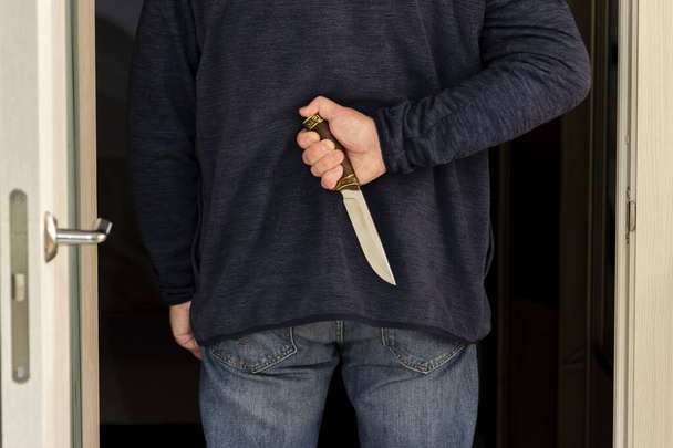 Un uomo entra dalla porta di una stanza buia e tiene un coltello in mano dietro la schiena. - Foto, immagini