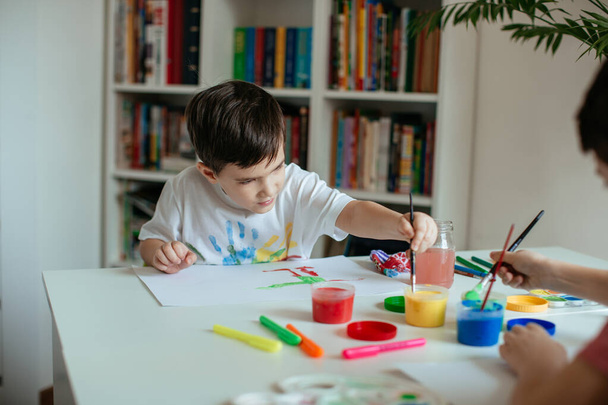 Αριστερό μικρό αγόρι που πάει να πάρει μπογιά με πινέλο στο χέρι. Παιδί με πινέλο και πολύχρωμα χρώματα στο σπίτι. - Φωτογραφία, εικόνα