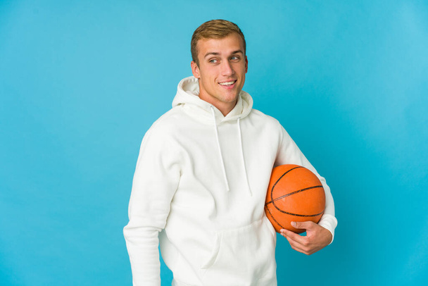 Nuori valkoihoinen mies pelaa koripalloa eristetty sinisellä taustalla näyttää syrjään hymyilevä, iloinen ja miellyttävä. - Valokuva, kuva