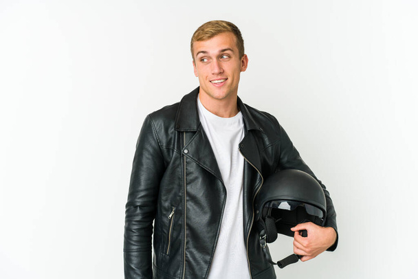 Joven hombre caucásico sosteniendo un casco de moto aislado sobre fondo blanco mira a un lado sonriente, alegre y agradable. - Foto, imagen