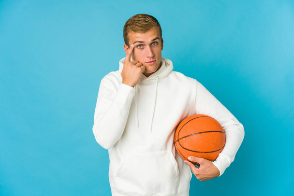 Giovane uomo caucasico giocare a basket isolato su sfondo blu cercando di ascoltare un pettegolezzo. - Foto, immagini