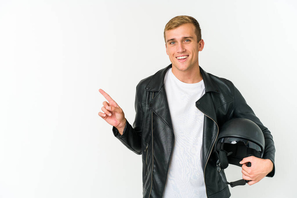Joven hombre caucásico sosteniendo un casco de moto aislado sobre fondo blanco sonriendo y señalando a un lado, mostrando algo en espacio en blanco. - Foto, Imagen