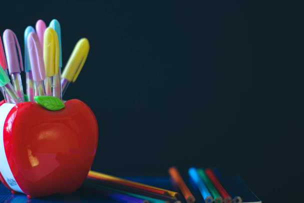 School accessories. Multicolored Pencils Stand at the stands in the shape of an apple. In Background of a dark school board and a piece of chalk. On the board is a place for a copy space. - Photo, Image