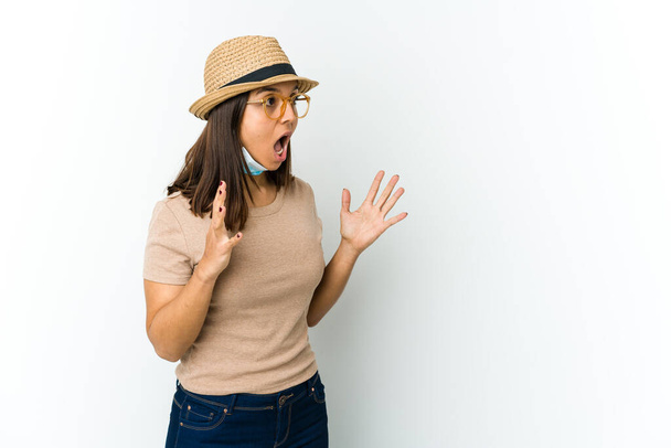 Jovem mulher latina usando chapéu e máscara para proteger de covid isolado no fundo branco gritos em voz alta, mantém os olhos abertos e as mãos tensas. - Foto, Imagem