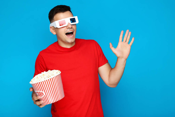 Young man with bucket of popcorn and eyeglasses on blue background - 写真・画像