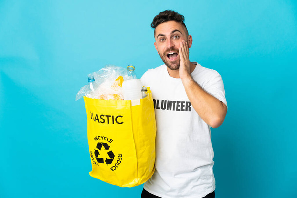 Белый мужчина держит сумку, полную пластиковых бутылок для переработки изолированы на синем фоне кричать с открытым ртом - Фото, изображение