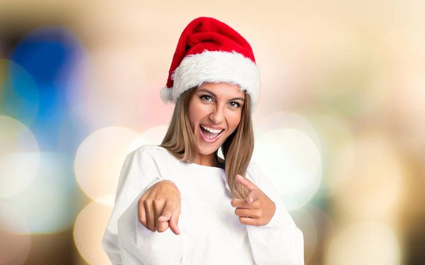 Girl with christmas hat points finger at you over unfocused background - Photo, Image