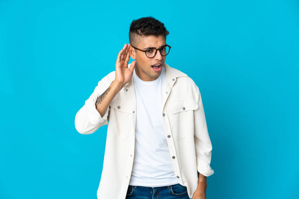 Young Brazilian man isolated on blue background listening to something by putting hand on the ear - Photo, Image