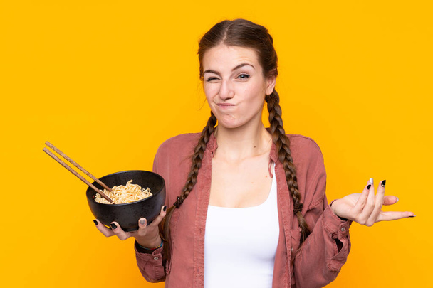 Young woman over isolated yellow background making doubts gesture while lifting the shoulders while holding a bowl of noodles with chopsticks - Фото, изображение