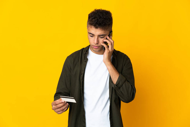 Nuori brasilialainen mies eristetty keltaisella taustalla ostaa matkapuhelimella luottokortilla - Valokuva, kuva