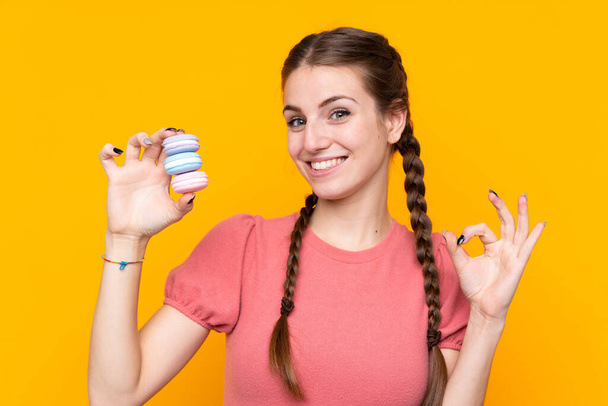 Молода жінка на жовтому фоні тримає барвисті французькі макарони і показує пальцями знак. - Фото, зображення