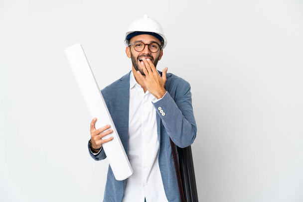 Young architect man with helmet and holding blueprints isolated on white background happy and smiling covering mouth with hand - Photo, Image