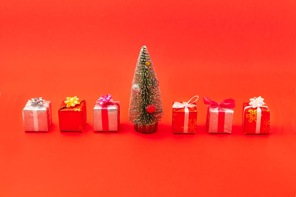 Nieuwjaarssamenstelling. Groene kerstboom met decoraties en dozen met geschenken gebonden met kleurrijke linten met strikken. Isoleer op rode achtergrond - Foto, afbeelding