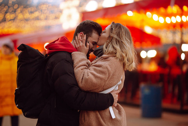 Happy couple in love walking on Christmas market at evening. Young woman and man at festive street market  enjoying winter moments. Lights around. Christmas, New Year. - Foto, afbeelding