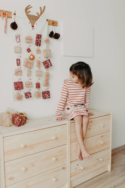 Cute girl opening Christmas Advent calendar gifts. Wrapped gifts for children. Seasonal tradition. Eco friendly Christmas - 写真・画像