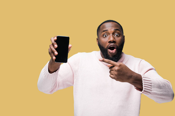Afro-Amerikaanse man in casual kleding staat op geïsoleerde oranje achtergrond en wijst een vinger naar de telefoon in verrassing terwijl hij naar de camera kijkt - Foto, afbeelding