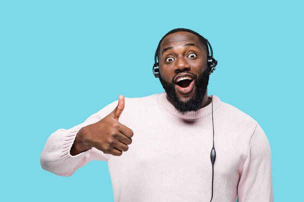 Portrait of cheerful african american call center worker in headphones with microphone standing on isolated blue background, surprised looks at camera and showing thumb up - Photo, Image