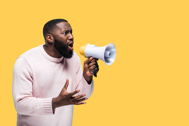 Podekscytowany czarny mężczyzna krzyczy głośno do megafonu jakieś ogłoszenie stojące na odizolowanym żółtym tle - Zdjęcie, obraz
