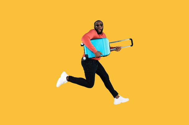 Zufriedener afroamerikanischer Mann mit Sonnenbrille und Freizeitkleidung, der mit einem blauen Koffer in der Hand auf orangefarbenem Hintergrund springt. In Vorfreude auf die Reise - Foto, Bild