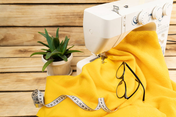 Máquina de coser blanca con tela amarilla, cinta de centímetro y vasos, en el fondo una flor en macetas blancas, sobre un fondo de madera. Concepto de negocio doméstico. - Foto, imagen
