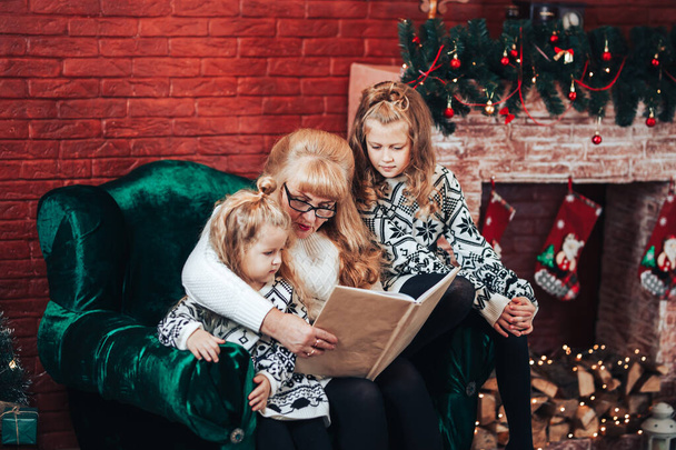 Großmutter mit Kindern las, ein Buch in der Hand haltend. Weihnachtsstimmung. Im Hintergrund gibt es einen Kamin und einen grünen Sessel. - Foto, Bild