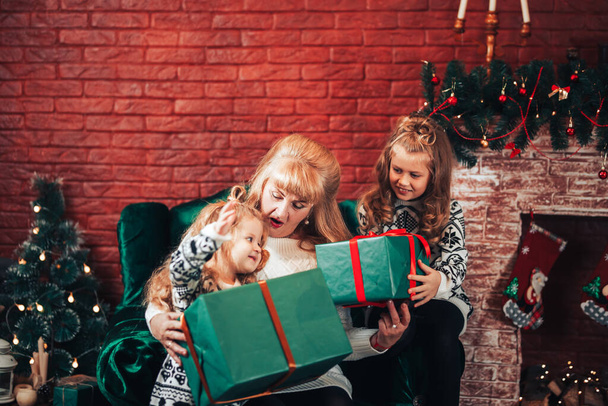 Babcia daje dzieciom prezenty świąteczne w zielonym opakowaniu. Boże Narodzenie. W tle znajduje się kominek i zielony fotel. - Zdjęcie, obraz