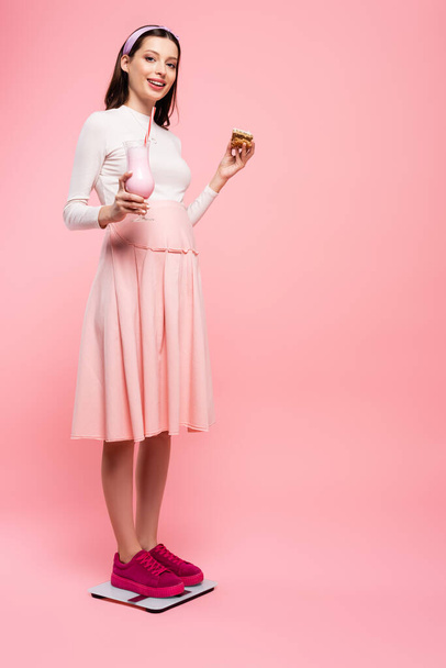giovane bella donna incinta con torta e frullato su scale isolate su rosa - Foto, immagini