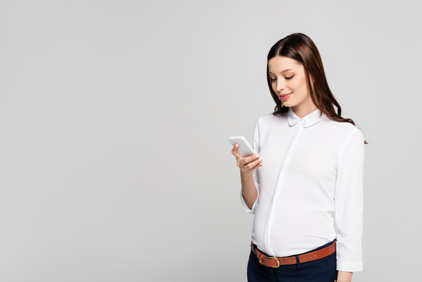 sonriente joven embarazada mujer de negocios con teléfono inteligente aislado en gris - Foto, imagen