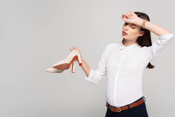 tired young pregnant businesswoman holding heeled shoes isolated on grey - Photo, Image