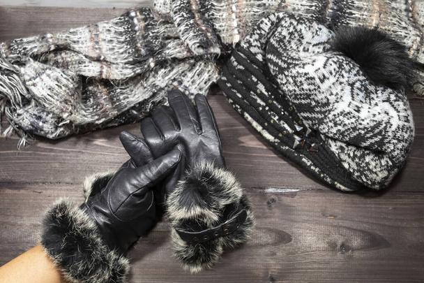 Auf dunklem Hintergrund liegen eine Wollmütze und ein Schal. Eine Hand in einem schwarzen Lederhandschuh mit Fell hält einen zweiten Handschuh. - Foto, Bild